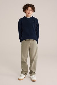 Marinblå stickad tröja med vit krage, beige löst sittande byxor och vita sneakers med gummisulor. Modellen står mot en neutral bakgrund.