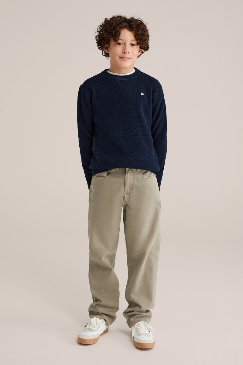 Marinblå stickad tröja med vit krage, beige löst sittande byxor och vita sneakers med gummisulor. Modellen står mot en neutral bakgrund.