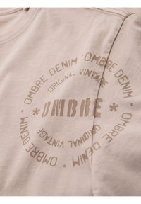 Béžové bavlněné tričko s kulatým grafickým potiskem v barvě pučálky, s nápisy "OMBRE DENIM" a "ORIGINAL VINTAGE". Měkká struktura, krátké rukávy.