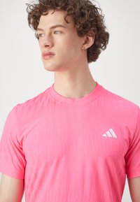 Pink athletic shirt with a textured geometric pattern, round neckline, short sleeves, and white Adidas logo on the upper left side.