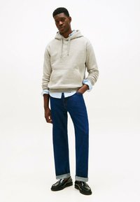 Giovane uomo che indossa un felpa grigia con cappuccio, una camicia blu a bottoni, jeans blu scuri con risvolto e mocassini neri, in piedi con una mano in tasca.