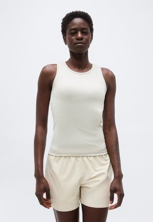 Atletska oseba, oblečena v svetlo bež brezrokavnik in ujemajoče se kratke hlače, stoji z rokami sproščenimi ob telesu na enobarvni beli podlagi.