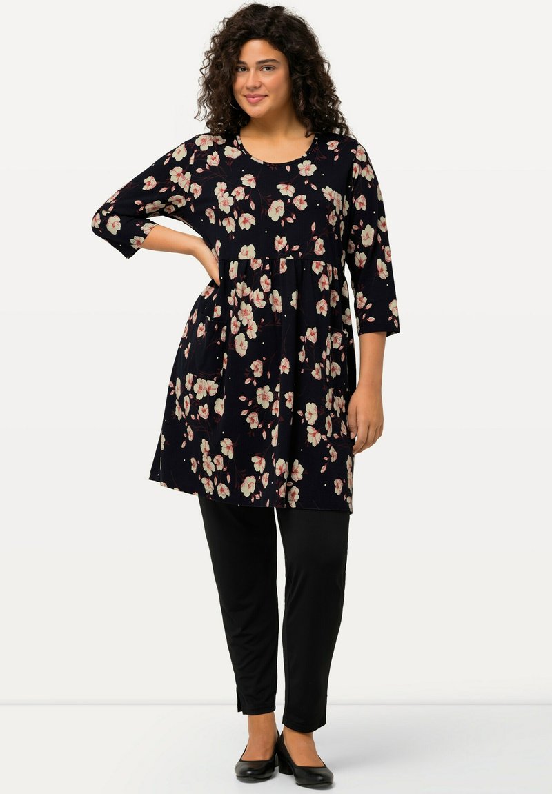 Florale Tunika mit schwarzem Hintergrund, rundem Ausschnitt, dreiviertellangen Ärmeln, kombiniert mit schwarzer Hose und flachen Schuhen.