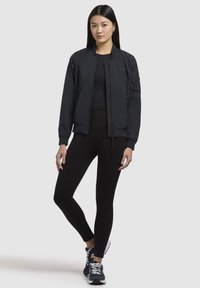 Giacca bomber scura con zip frontale, polsini a coste e tasca con zip sulla manica, abbinata a leggings neri e scarpe da ginnastica blu.