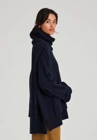 Kvinna iförd en överdimensionerad marinblå polotröja kombinerad med senapsgula byxor, stående mot en enfärgad grå bakgrund, sedd från sidan.