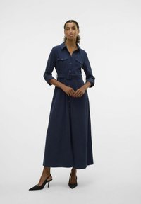 Robe chemise longue en navy avec un devant à boutons, des manches retroussables, deux poches poitrine et une ceinture assortie à la taille. Escarpins noirs à bride arrière.