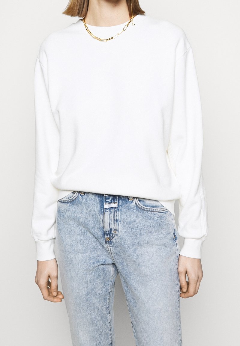 Personne portant un sweatshirt blanc à manches longues, un jean taille haute bleu clair et un collier en chaîne dorée sur un fond uni.