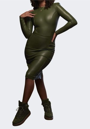 Vrouw draagt een aansluitende, olijfgroene leren jurk met lange mouwen en bijpassende veterlaarsjes tot aan de enkels, poseert met één hand op haar heup en de andere bij haar borst.