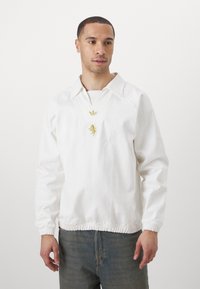 Bílá pulovrová bunda s límečkem, žebrovanými manžetami a elastickým lemem. Obsahuje zlaté logo na hrudi.
