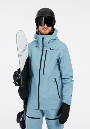 Helleblaue wasserdichte Snowboardjacke mit schwarzen Reißverschlüssen und Akzenten, kombiniert mit passenden Hosen und schwarzen Handschuhen. Snowboard wird in einer Hand gehalten.