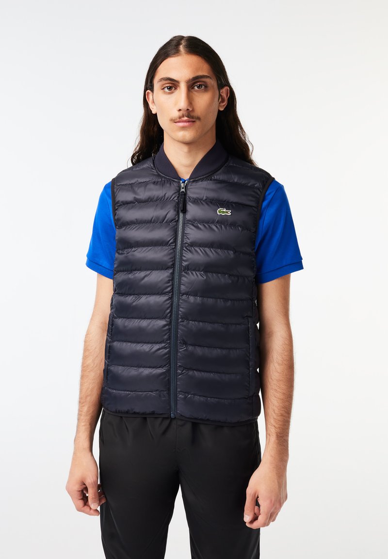 Gilet matelassé navy avec fermeture éclair à l'avant, motif cousu horizontalement et col côtelé. Présente un petit logo vert sur la poitrine.