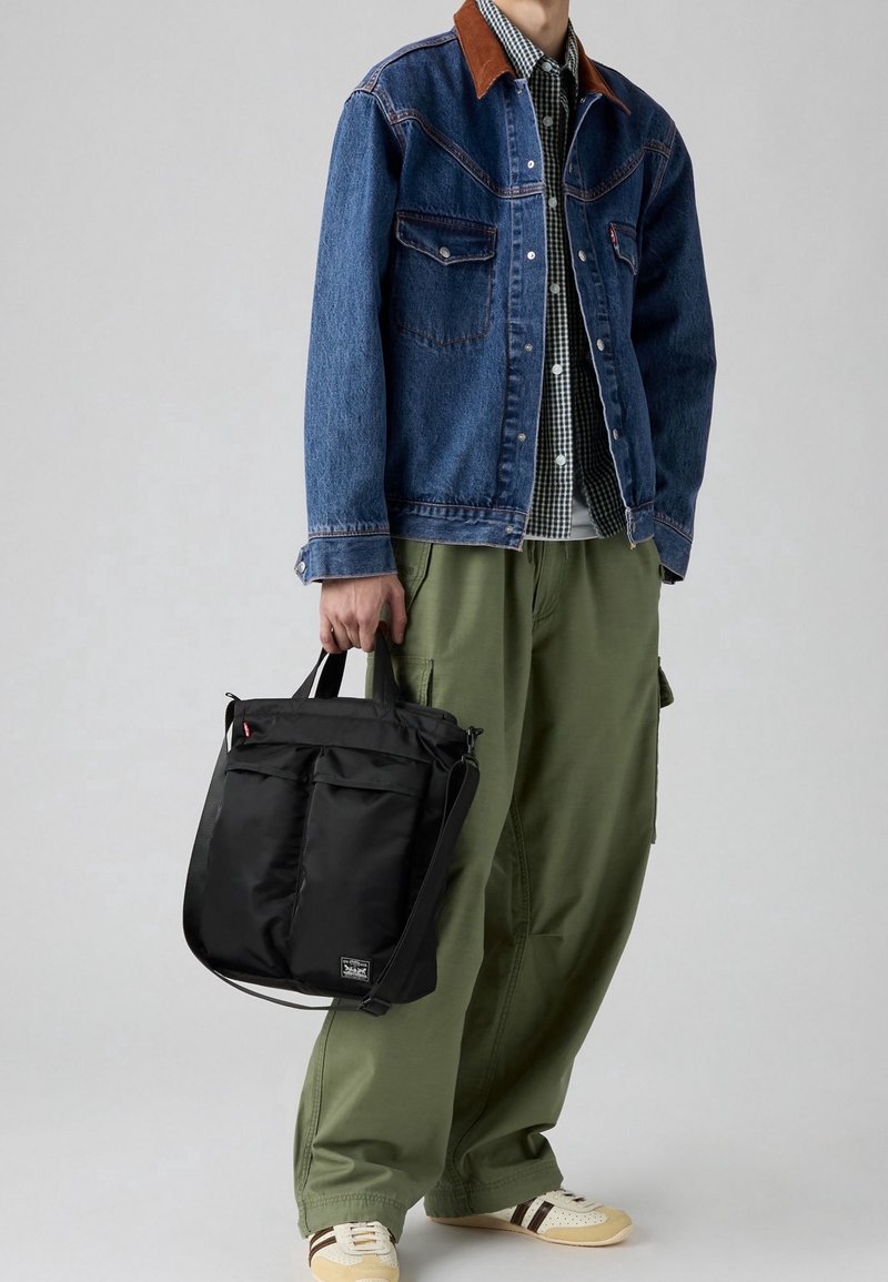 Personne portant une veste en jean bleue, un pantalon vert à jambes larges, des baskets beiges, tenant un sac à bandoulière noir avec double poignée et poches avant.