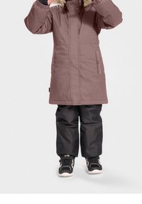 Manteau marron isolé avec capuche en fourrure, comportant des poches avant et des zips. Associé à un pantalon de neige noir imperméable et des baskets noires.