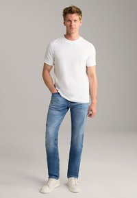 T-shirt bianca a maniche corte, jeans azzurri con effetto sbiadito e sneaker bianche. Il modello sta in piedi con una mano in tasca su uno sfondo neutro.