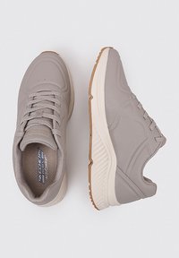 Zapatillas grises con un upper sintético suave, diseño con cordones y plantillas acolchadas. Presenta una mediasuela de color crema contrastante y suela de goma.