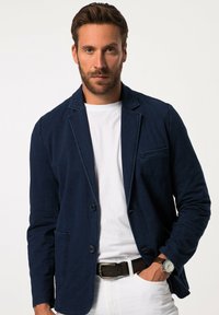 Marineblå blazer i ett mjukt tyg, enkelknäppt med slagkrage, framficka och två knappar. Bärs över en vit skjorta.