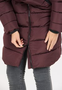 Burgundy pufferjacka med horisontella sömmar, svart dragkedja och elastiska ärmslut. Visar en hand som justerar dragkedjan vid jackans nederkant.