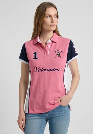 Mujer con un polo rosa y azul marino con los logotipos "1" y "Valecuatro", de pie con una mano en el bolsillo y mirando de lado.
