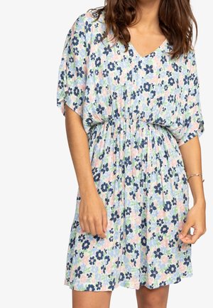 Kvinde iført en kort blomsterkjole med marineblå, lyserøde og lyseblå blomster samt elastisk talje, stående med armene afslappet langs siden.