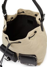 Tan canvas bucket bag with black leather accents, drawstring closure, and external zippered pocket. Smooth texture, structured shape.