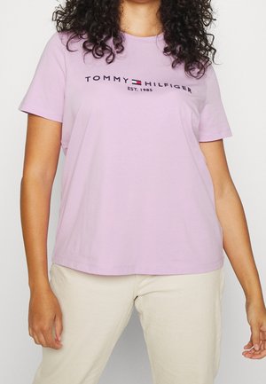 Žena nosící světle fialové tričko Tommy Hilfiger s krátkým rukávem a logem a béžové kalhoty, kudrnaté vlasy viditelné od ramen dolů.