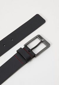 Ceinture en cuir noir avec une boucle rectangulaire en métal argenté ornée d'un logo en relief, des coutures rouges et une texture lisse.