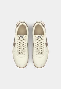 Zapatillas deportivas de cuero beige con detalles texturizados, punta redonda y una marca de color marrón claro. Cuentan con cordones planos y suela de goma para tracción.