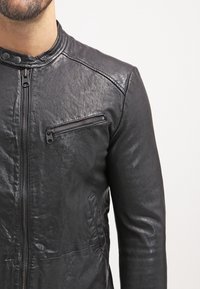 Uomo che indossa una giacca di pelle nera con zip frontale, tasca sul petto con zip e colletto con bottoni a scatto, su uno sfondo bianco semplice.