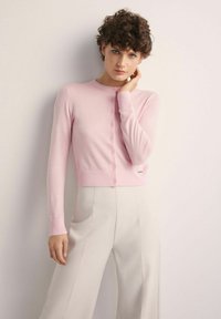 Młoda kobieta z kręconymi włosami nosi jasnoróżowy sweter zapinany na guziki oraz jasnobeżowe spodnie z wysokim stanem, stojąc przy jasnej ścianie.