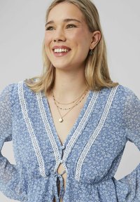 Blouse fleurie bleue avec des manches longues en voile, bordée de dentelle blanche, et un design à nouer à l'avant. Elle présente un tissu léger et texturé, ainsi que des colliers superposés.