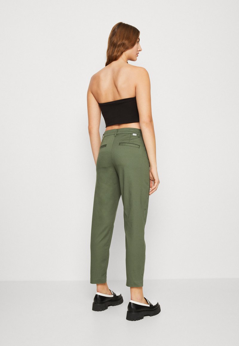 Pantalons verts fuselés avec deux poches arrière, associés à un haut noir sans bretelles et des chaussures noires avec des accents blancs. Texture de tissu lisse.