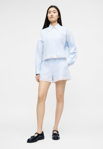 Νέα γυναίκα φορώντας ανοιχτό μπλε σετ με μακρυμάνικη μπλούζα και σορτς με λευκή δαντέλα, σε συνδυασμό με μαύρα παπούτσια Mary Jane, στέκεται σε λευκό φόντο.