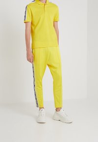Polo jaune à manches courtes avec col, assorti à un pantalon jaune avec des bandes noires sur les côtés. Des baskets blanches complètent la tenue.