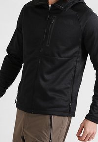 Zwarte ritssluiting hoodie gemaakt van zachte fleece met een textuurafwerking, voorzien van een borstzak aan de voorkant, zijzakken en een capuchon.