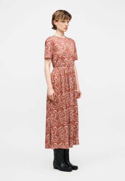 Γυναίκα με κοντά μαλλιά, ντυμένη με midi φόρεμα με κόκκινο και άσπρο λουλουδάτο σχέδιο και μαύρες μπότες μέχρι τη μέση της γάμπας, στέκεται μπροστά σε απλό φόντο.