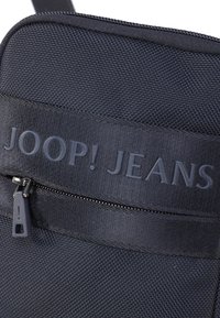 Tmavě modrá textilní taška na rameno s texturovaným povrchem, se zipovým uzávěrem a embosovaným logem "JOOP! JEANS" na přední straně.