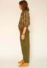 Chemise à carreaux dans des tons de marron et de jaune, pantalons cargo verts avec poches latérales, et baskets en daim jaunes avec des rayures brunes.