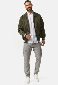 Giacca bomber verde oliva con accenti a strisce bianche, indossata sopra una camicia bianca a bottoni, abbinata a pantaloni cargo grigio chiaro e sneakers bianche.