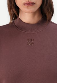 Brun høyhalset topp laget av glatt stoff. Har ribbet halslinning og brodert "HUGO"-logo i en mørkere brunfarge.