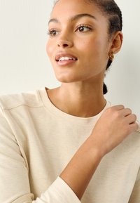 Kermanvärinen collegepaita, jossa teksturoitu kangas ja pyöreä pääntie. Malli käyttää kultaisia rengaskorvakoruja ja esittelee luonnollisen meikkityylin.