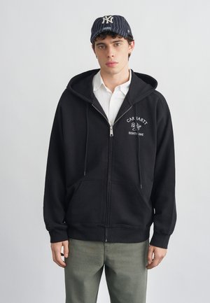 Giovane uomo che indossa una felpa nera con zip e la scritta "Carhartt", maglietta bianca, pantaloni color oliva e un cappellino da baseball a righe scure con un logo bianco.