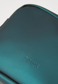 Borsa impermeabile color turchese con finitura liscia. Presenta il logo "RAINS" in rilievo, bordi arrotondati e design elegante, adatta a vari utilizzi.