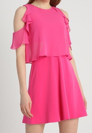 Femme portant une robe rose vif à épaules dénudées avec des manches à volants et un corsage à couches, posant devant un fond uni.