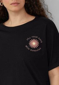 Zwarte katoenen T-shirt met ronde hals en korte mouwen. Beschikt over een roze madeliefje grafiek en de tekst "VUL JE DAGEN MET ZONNESCHIJN."