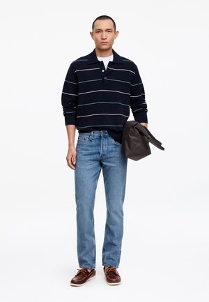 Homme portant un pull rayé bleu marine, un jean bleu clair et des chaussures marron, tenant un sac zippé marron foncé dans une main.