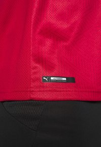 Punainen urheiluvaate, jossa on teksturoitu pinta, Puma-logo ja musta kangasviimeistely. Mukana on tarra lisenssitiedoilla.