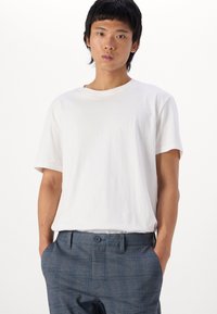 Weißes Baumwoll-T-Shirt mit Rundhalsausschnitt und kurzen Ärmeln, kombiniert mit grauen karierten Hosen, die über einen Knopf und Gürtelschlaufen verfügen.
