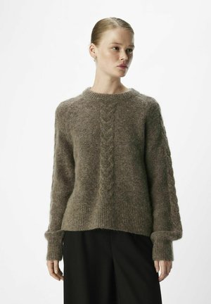 Bruine mohair trui met een kabelbreipatroon in het midden, geribbelde mouwen en een losse pasvorm, gecombineerd met zwarte broeken.
