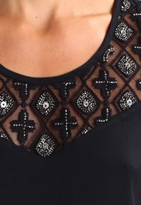 Top nero con scollatura trasparente, decorata con motivi a diamante e dettagli in perline metalliche. Tessuto liscio e design del colletto rotondo.