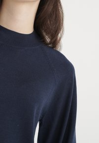 Granatowy dzianinowy sweter z wysokim kołnierzem, wykonany z tkaniny o fakturze i z rękawami raglanowymi, pokazany od szyi i okolicy ramion.